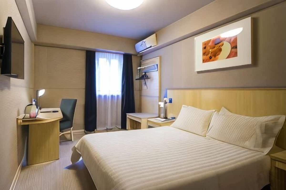 A hotel room with a large bed, a desk, a chair, a TV, and a window with curtains.