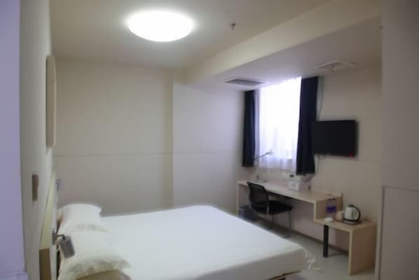 Quarto de hotel com uma cama de solteiro, uma mesa, uma cadeira, uma televisão presa à parede e uma janela com cortinas.