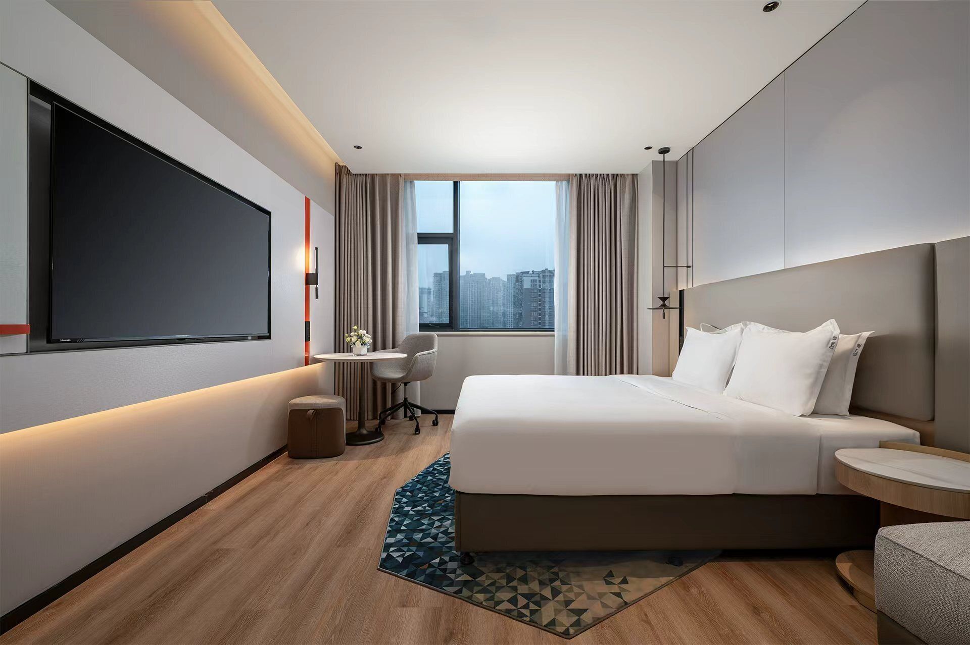 Una habitación de hotel moderna con una cama grande, un televisor de pantalla plana, una mesita con un jarrón y vistas a la ciudad por la ventana.