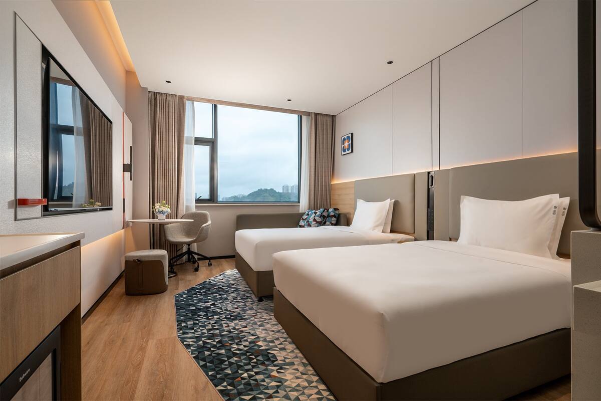 Une chambre d’hôtel avec deux lits, un bureau, une chaise, une petite table et une vue sur la ville.