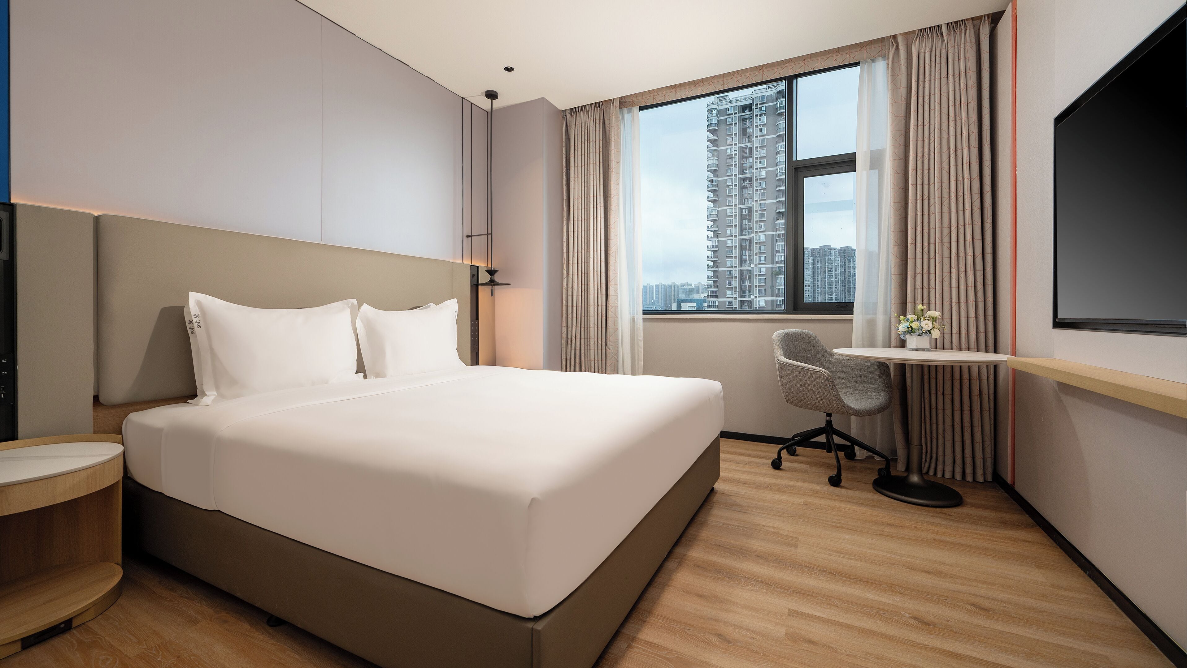 A hotel room with a large bed, a desk with a chair, a TV, and a window with a city view.