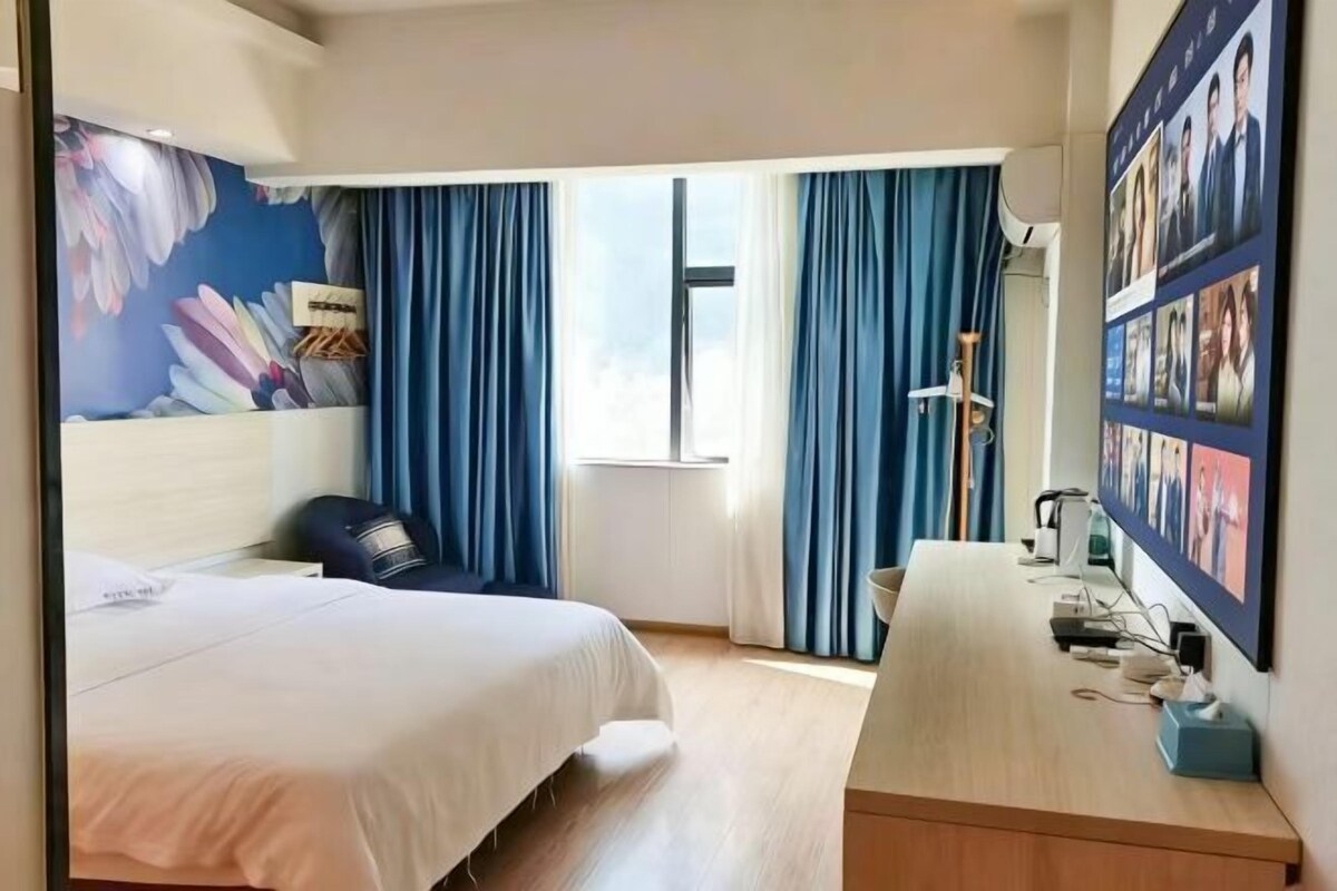 A hotel room with a bed, a desk, a chair, a window with curtains, a wall-mounted TV, and a wall with photos.