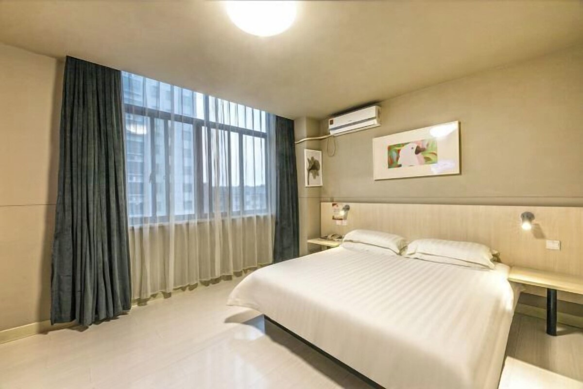 A hotel room with a large bed, a bedside table, an air conditioning unit, and a window with curtains.