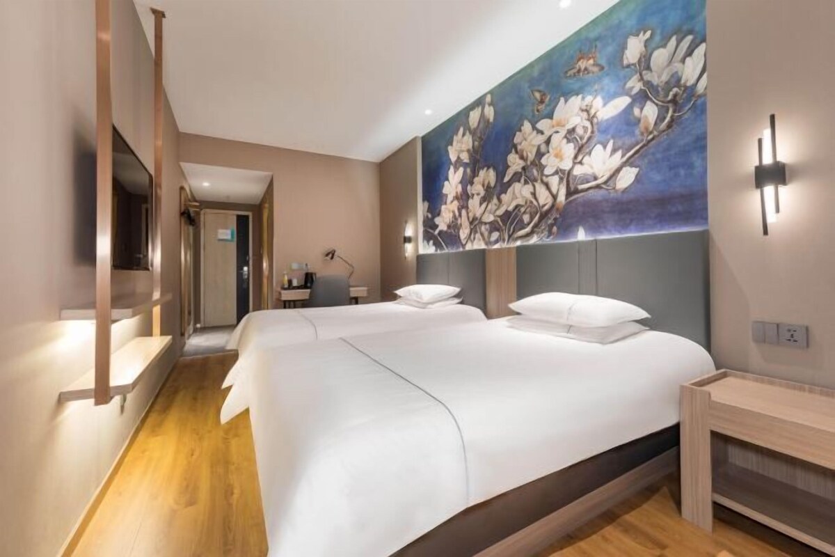 A hotel room with two beds, a wooden floor, a painting of flowers on the wall, and a desk with a chair.