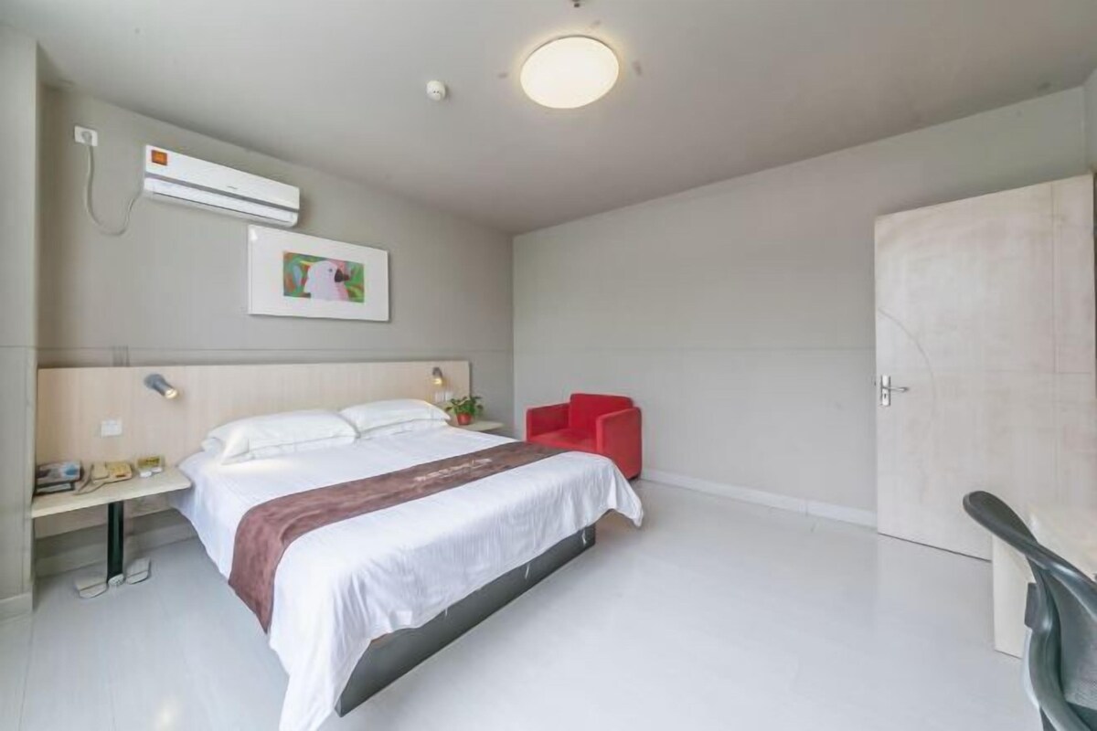 A modern hotel room with a bed, a desk with a chair, a red chair, an air conditioning unit, and a door.