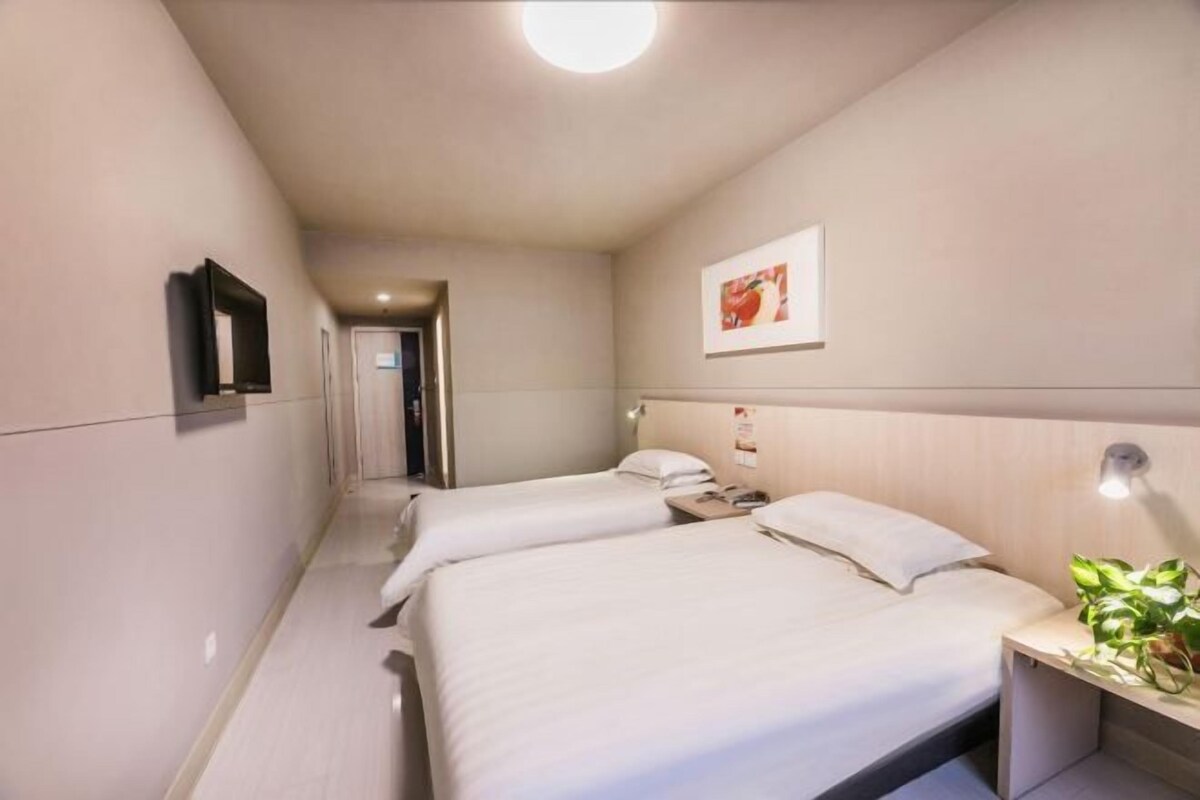 A hotel room with two beds, a flat-screen TV mounted on the wall, a bedside table with a lamp, and a framed picture on the wall.