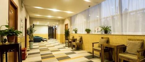 A hotel lobby with a checkered floor, seating area with chairs and tables, potted plants, and a large window with sheer curtains.