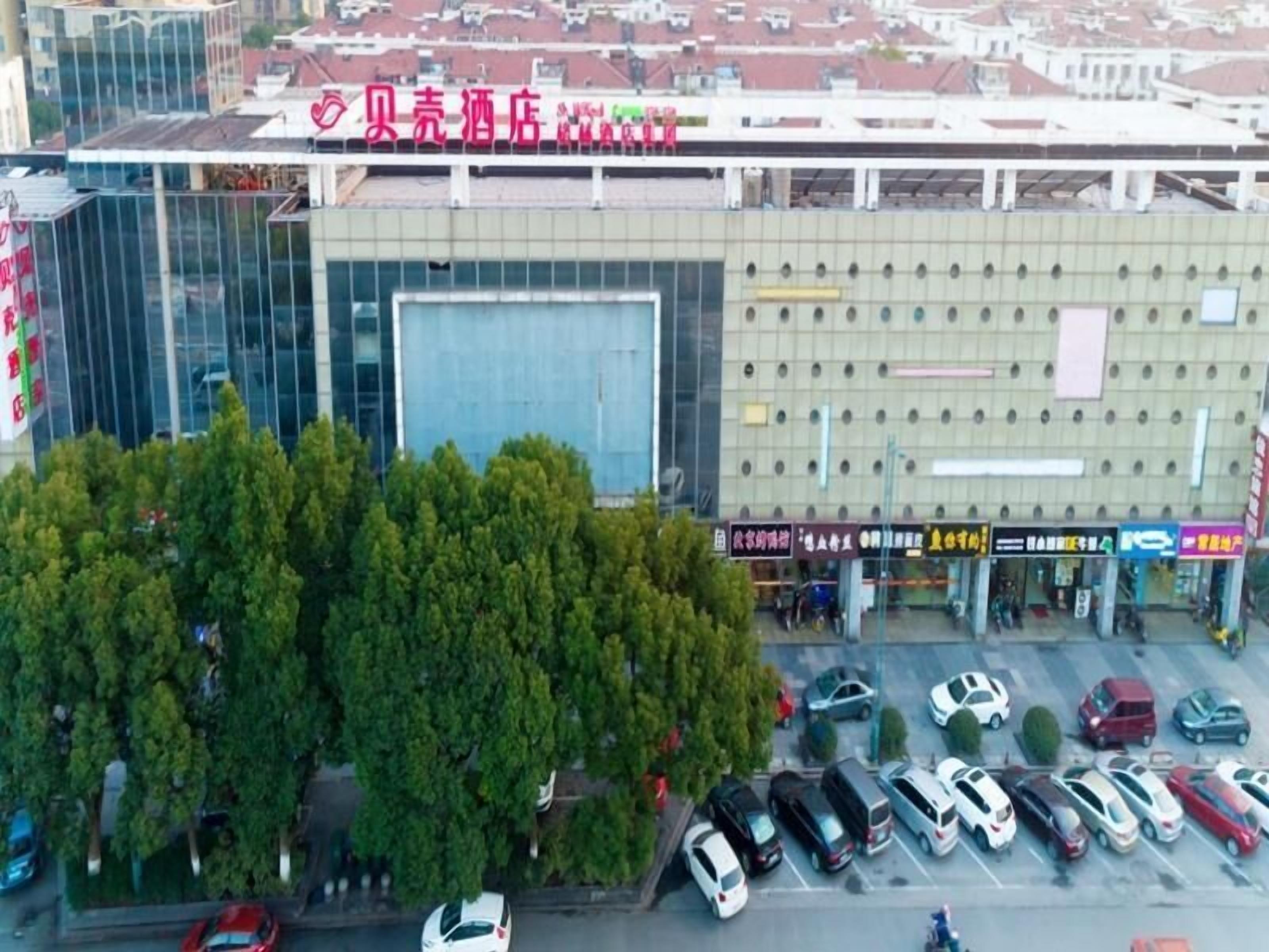 Ein Hotel mit moderner Glasfassade, ein Parkplatz mit mehreren Autos und eine Reihe von Geschäften mit fremdsprachlicher Beschilderung.