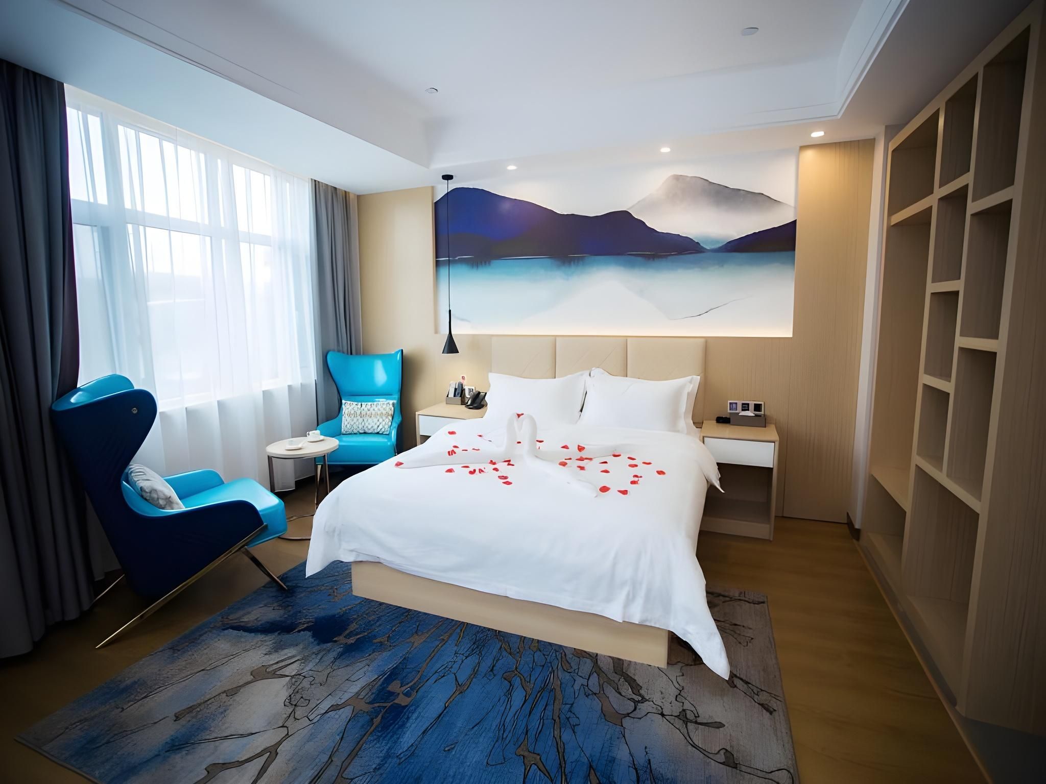 A hotel room with a bed, a blue armchair, a nightstand, a large painting of a landscape, and a wooden wardrobe.
