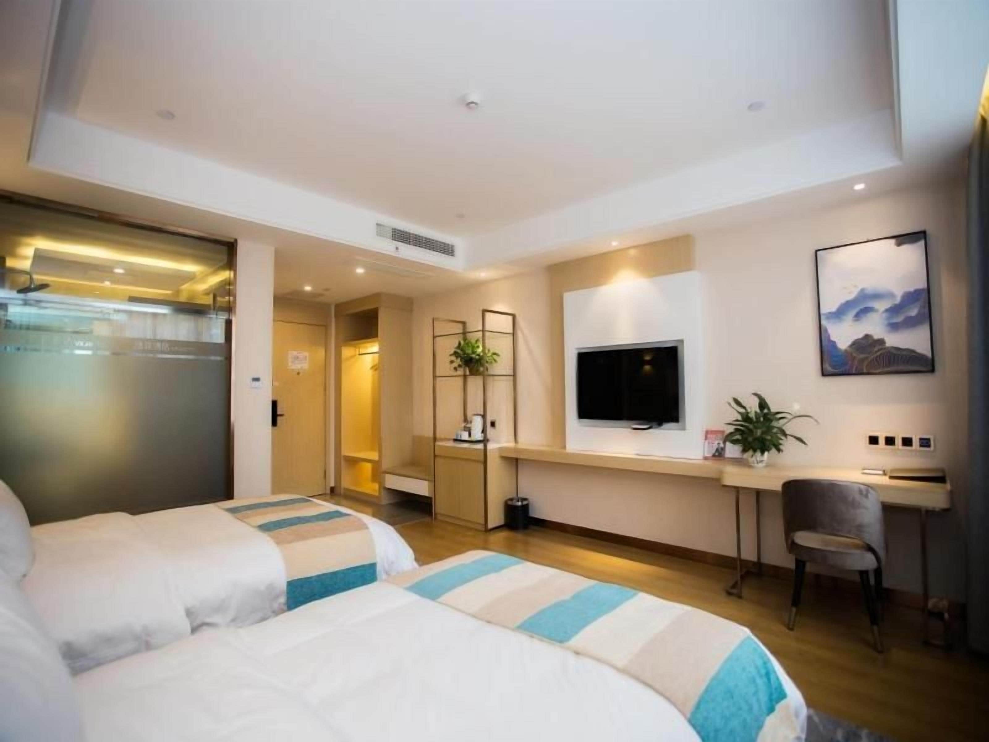 A hotel room with two beds, a desk, a chair, a TV, and a plant.