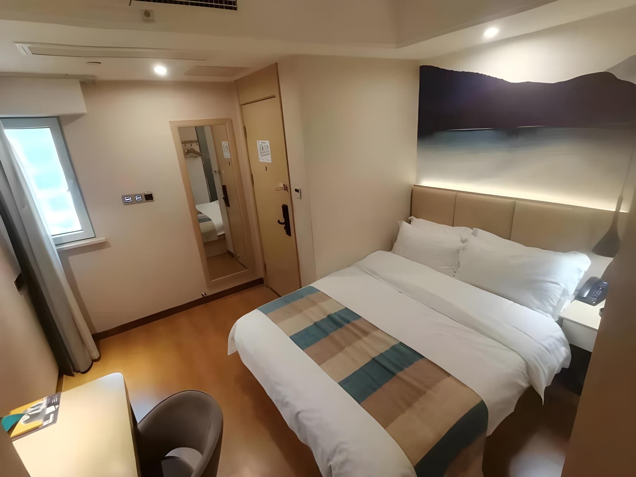 A hotel room with a bed, bedside table, chair, and a view of the hallway.