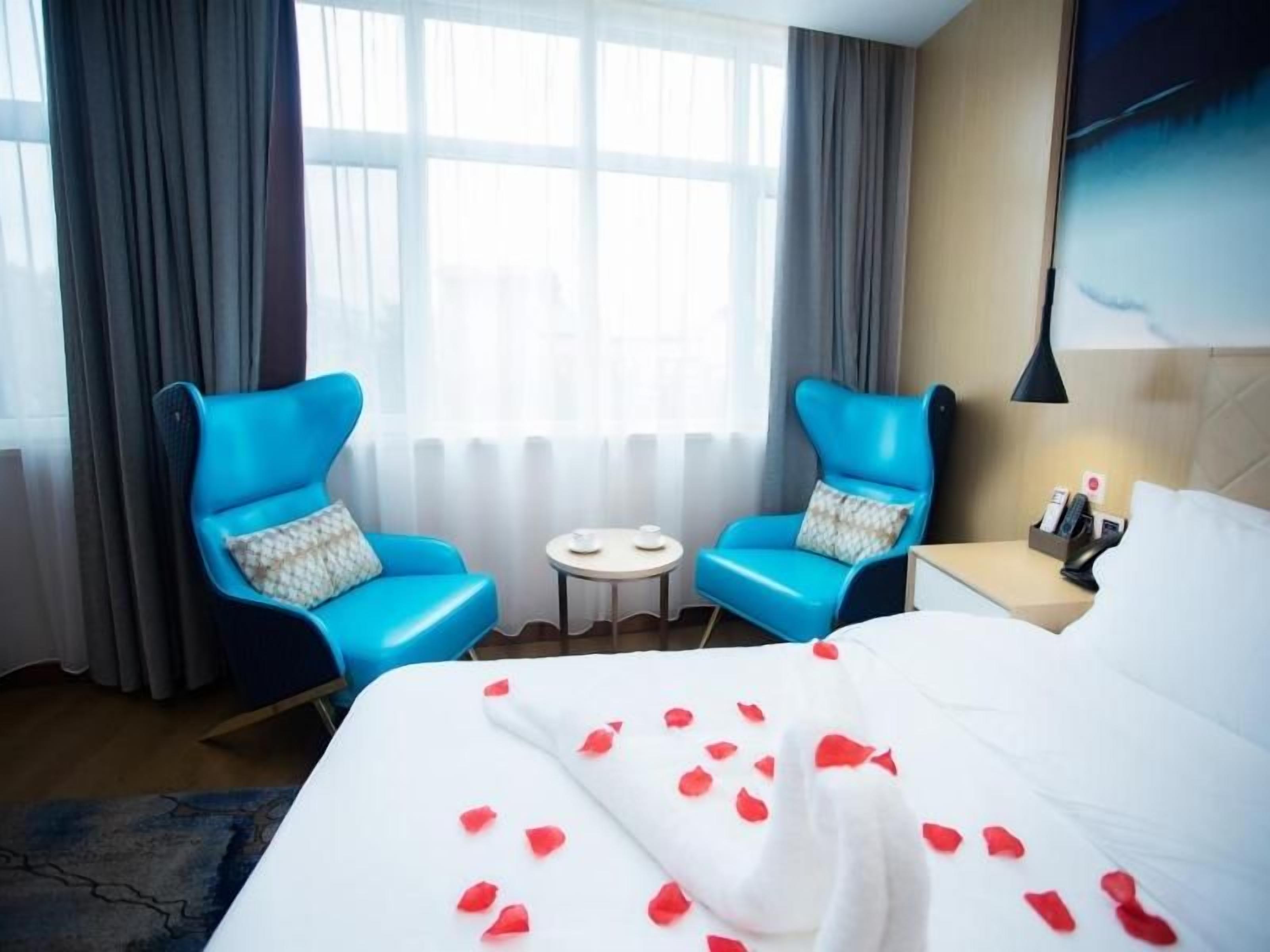 Habitación de hotel con una cama, dos sillones azules, una mesita y una mesita de noche con lámpara.