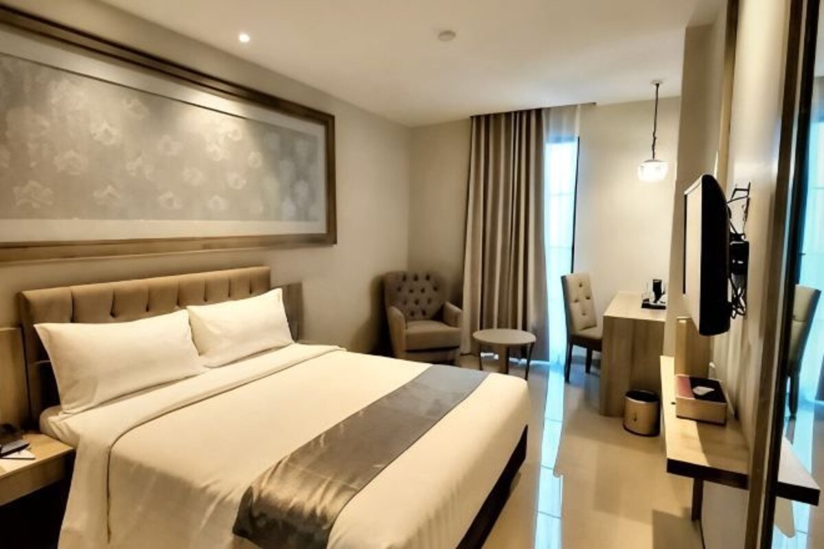 A modern hotel room with a large bed, a flat-screen TV, a seating area with a chair and a small table, and a large window with curtains.