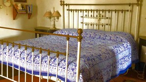 A bedroom with a metal bed frame, a floral bedspread, a nightstand with a lamp, and a wall-mounted shelf with books and decorative items. - Monte Alentejano T5 Private Pool (Portalegre)