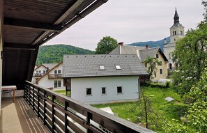 A traditional building with a sloped roof, a balcony with a wooden railing, and a view of a church with a tower in the background. - Apartments Znidar (Bohinj)