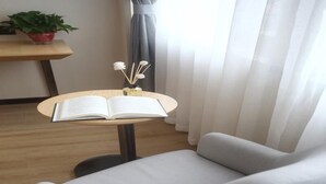A modern room with a white armchair, a small wooden table with an open book, a potted plant, and sheer curtains. - Greentree Inn Fuyang Exhibition Center (Fuyang)