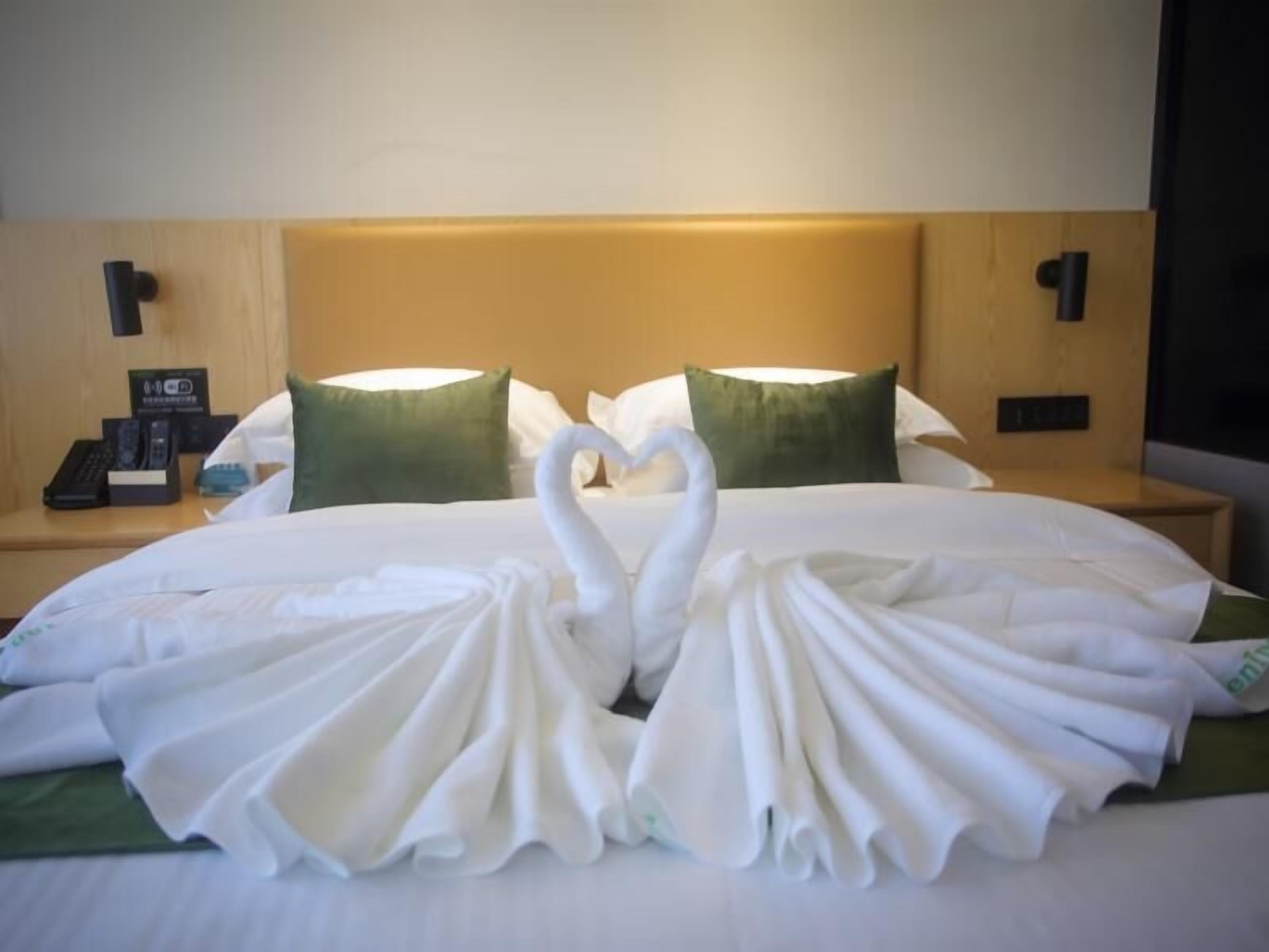 A hotel room with a neatly made bed, two green pillows, a bedside table with a phone and a card holder, and a wall-mounted light.