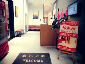 Zona de recepción de un hotel con mostrador, una banca, felpudo de bienvenida y cartel con tarifas de habitaciones.