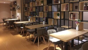 A modern interior with multiple wooden tables and chairs, a black metal shelving unit with various items, and a warm wooden floor. - 7 Days Premium (Chongqing Qijiang District Government Shop) (Guangzhou)