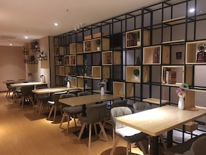 Un interior moderno con varias mesas y sillas de madera, una estantería de metal negro con diversos objetos, y un cálido suelo de madera.