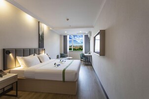 A hotel room with a bed, bedside table, lamp, and a view of the city through a window.