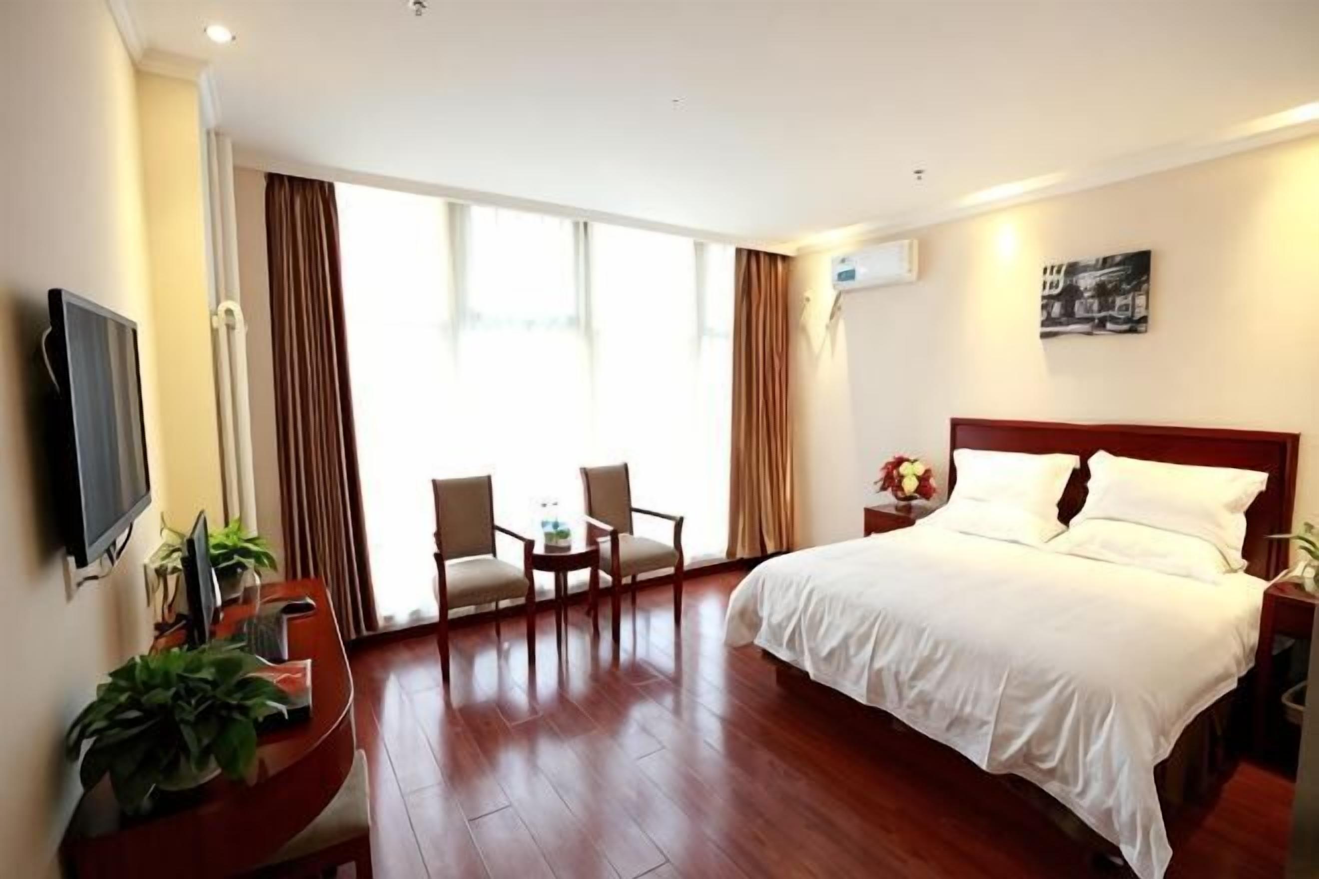 Quarto de hotel com uma cama, uma escrivaninha com cadeira, uma televisão e uma janela grande com cortinas.