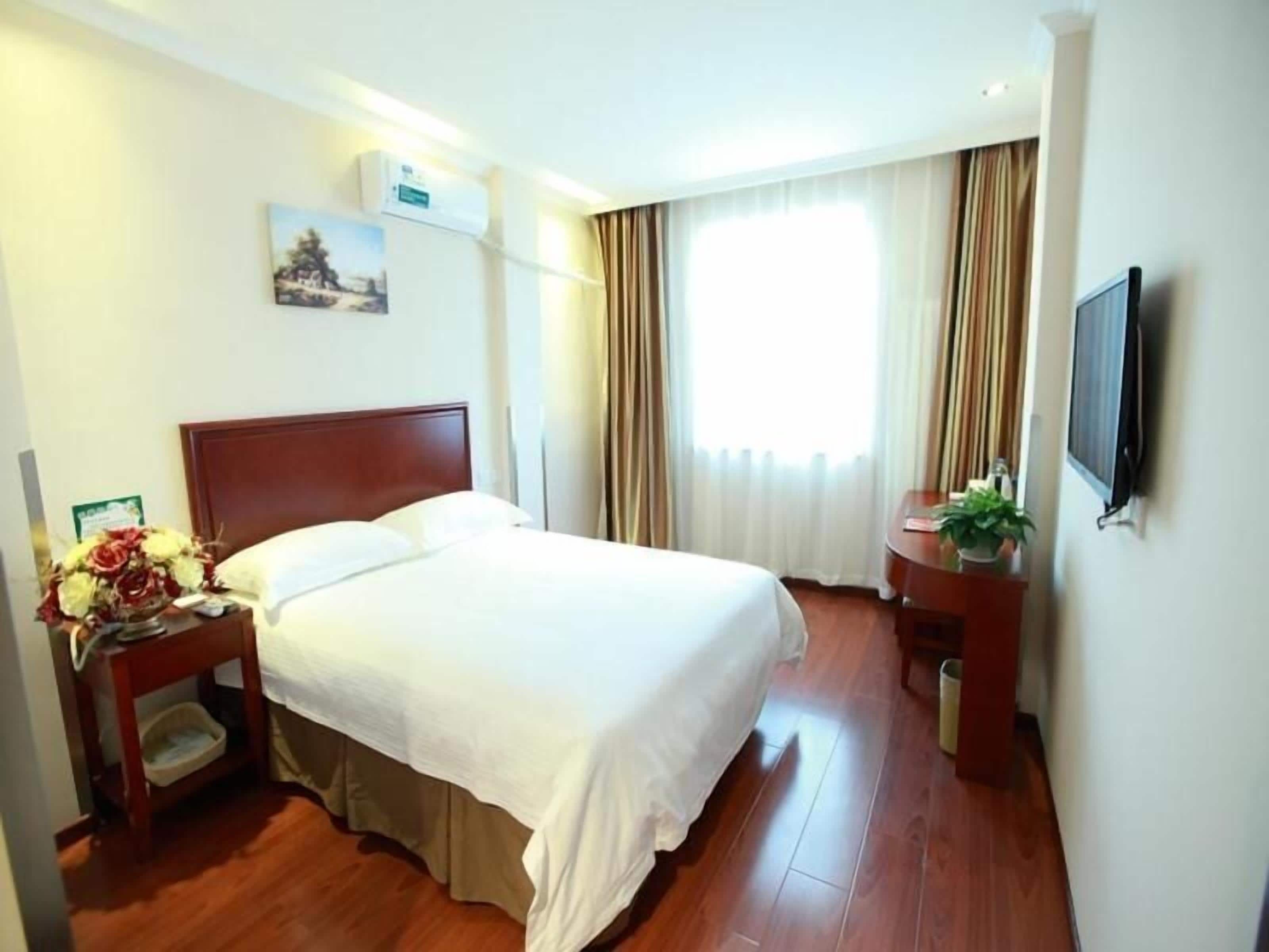 A hotel room with a wooden bed, a nightstand with a vase of flowers, a small table with a plant, a flat-screen TV, and a window with curtains.