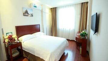 A hotel room with a wooden bed, a nightstand with a vase of flowers, a small table with a plant, a flat-screen TV, and a window with curtains.