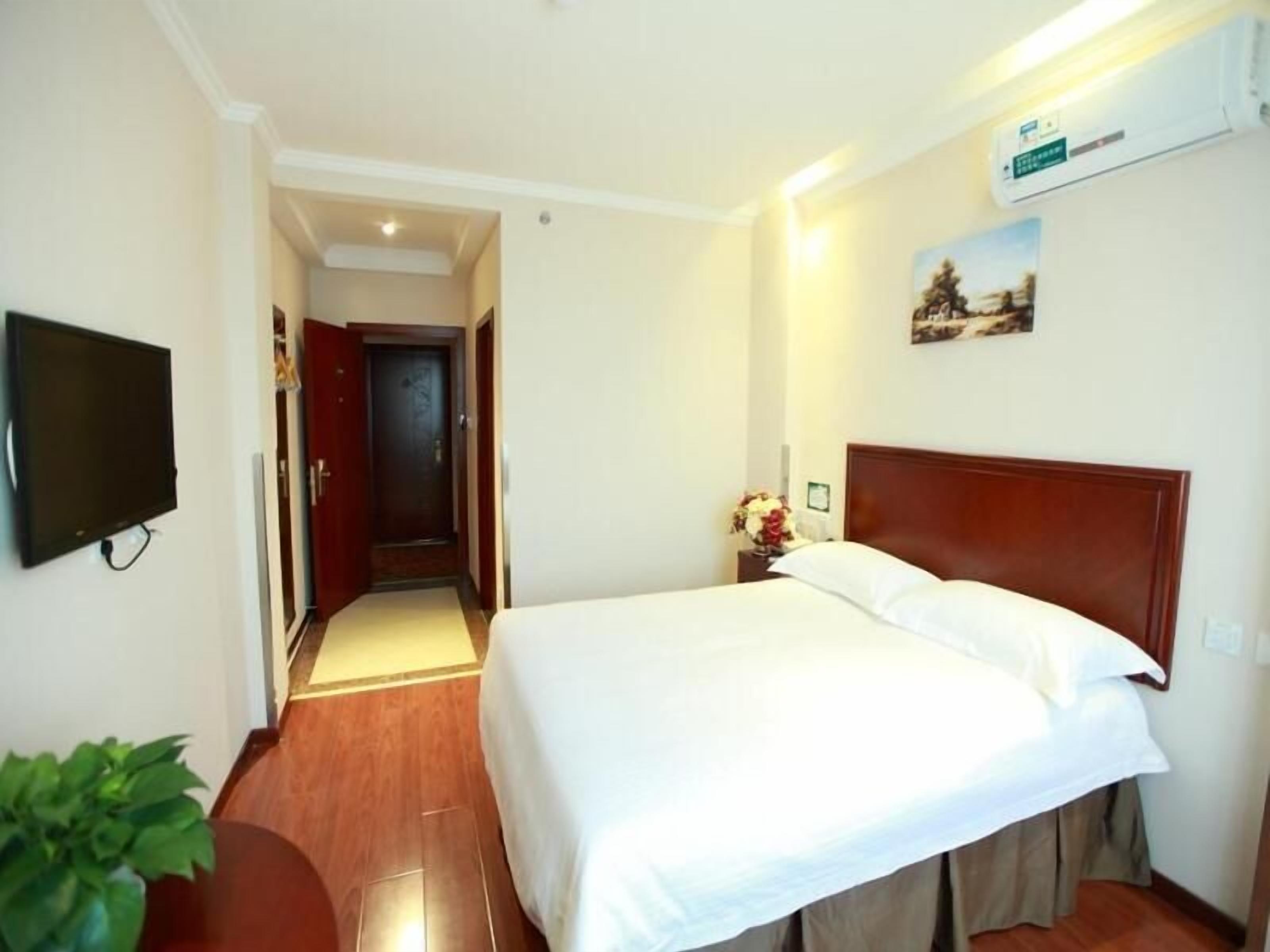 A hotel room with a bed, a TV, a wooden headboard, a nightstand with a vase of flowers, and a hallway leading to another room.