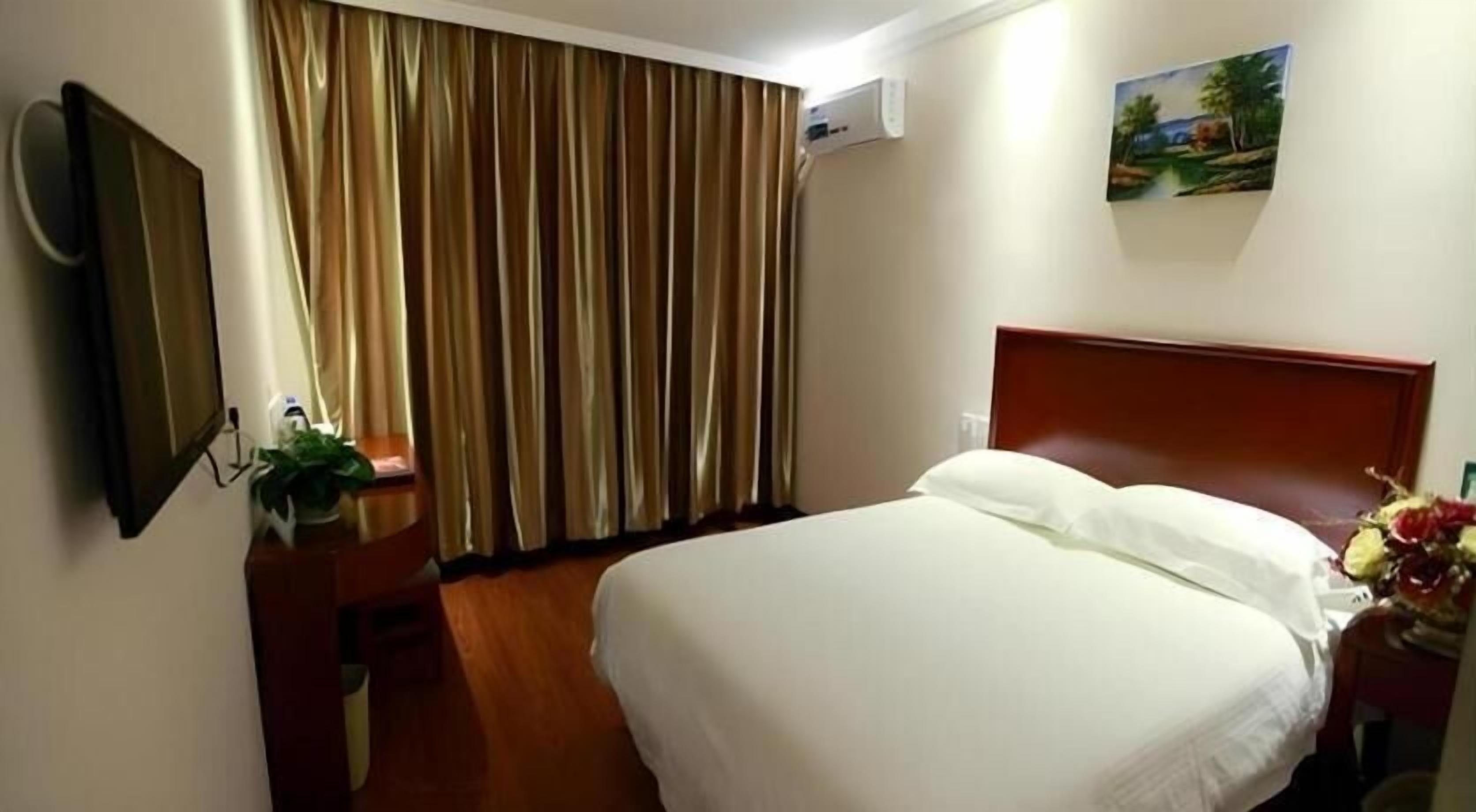 A hotel room with a bed, a wooden headboard, a nightstand with a plant, a mirror, a painting on the wall, and a window with curtains.