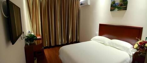 A hotel room with a bed, a wooden headboard, a nightstand with a plant, a mirror, a painting on the wall, and a window with curtains.
