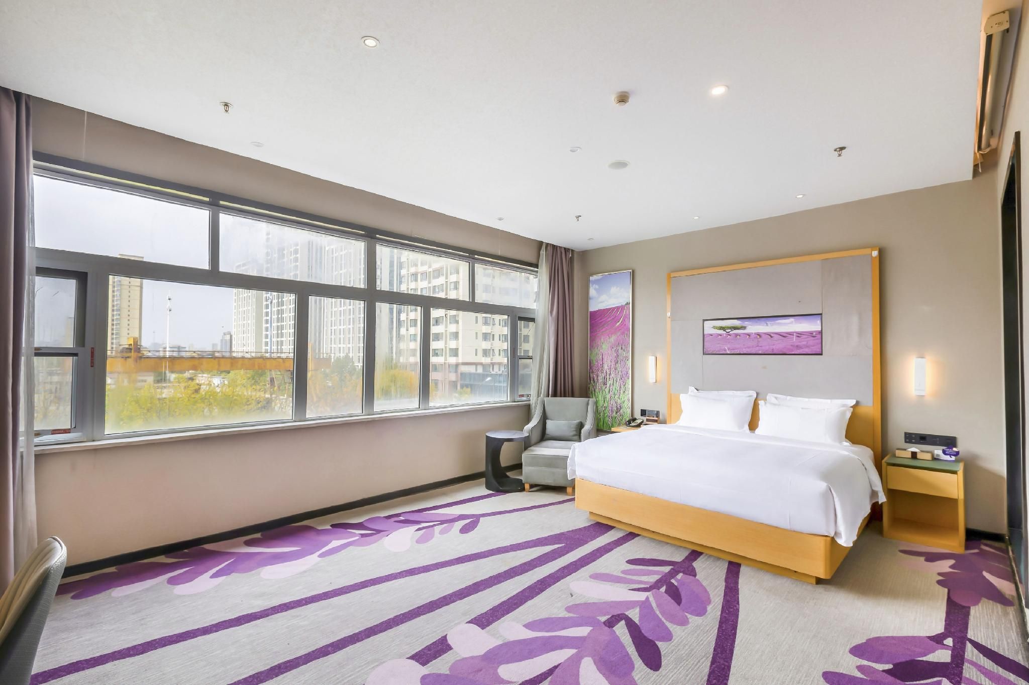 A hotel room with a large window, a bed, a chair, a bedside table, and a wall-mounted TV.