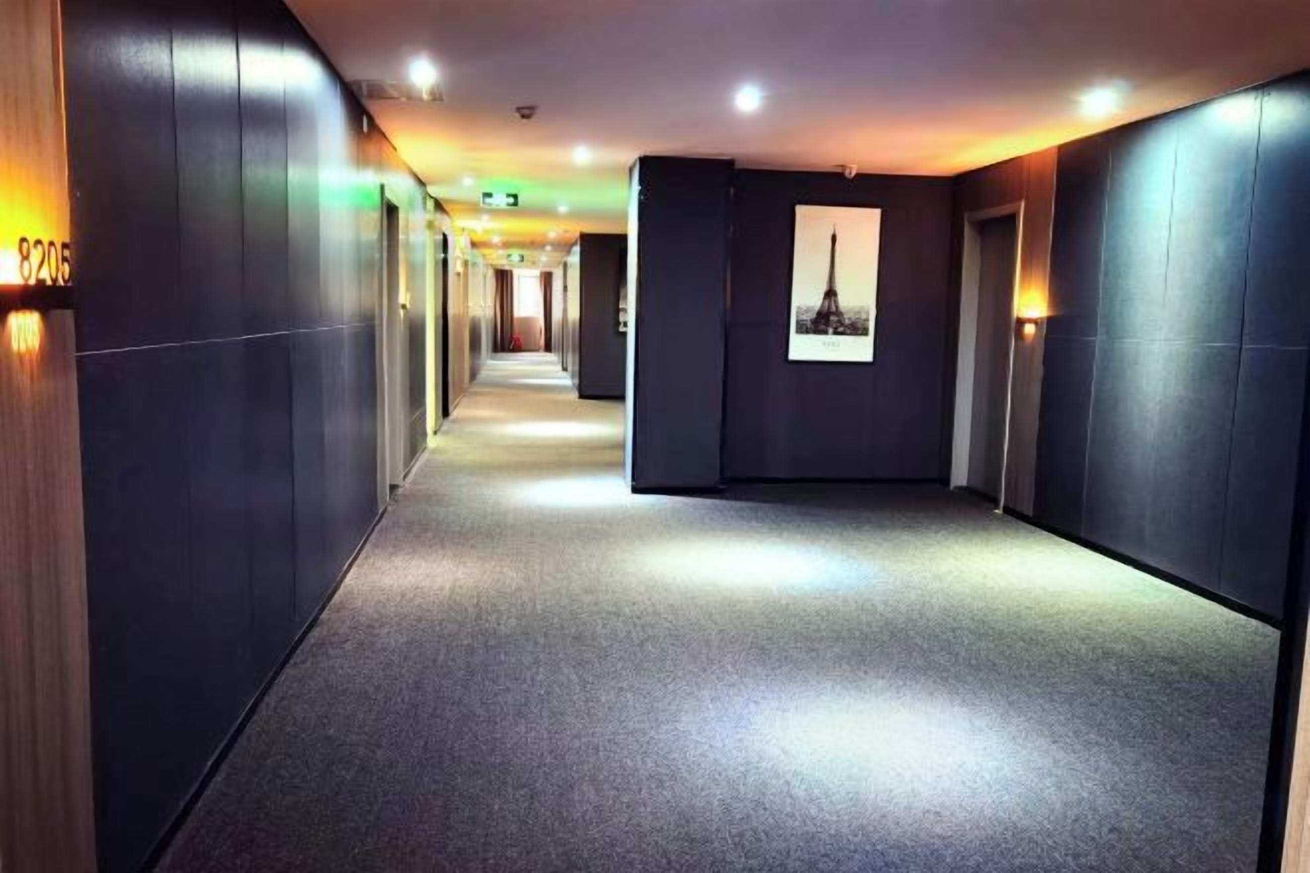 A hotel corridor with a carpeted floor, wall-mounted lights, and a framed picture on the wall.