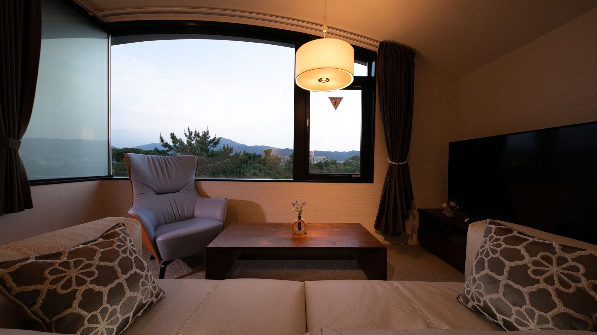 Ein Zimmer mit einer Couch, einem Sessel und einem Fernseher. Es gibt ein Fenster mit Blick auf Bäume und Berge.
