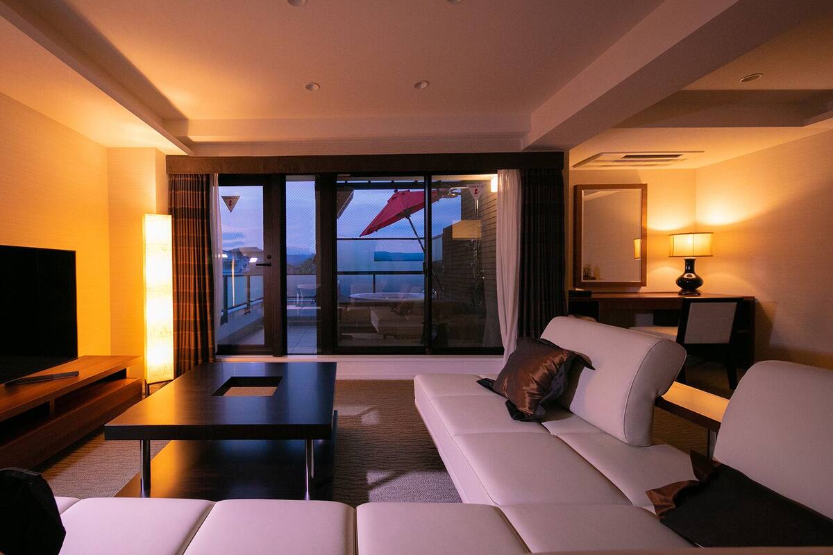 Une chambre d’hôtel équipée d’un canapé, d’une télévision, d’une table, d’une lampe et d’une porte-fenêtre coulissante donnant sur un espace extérieur.