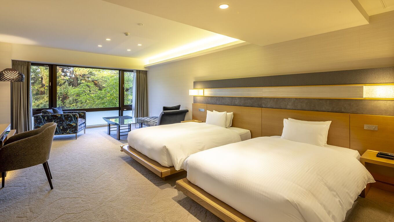 Ein Hotelzimmer mit zwei Betten, einem Schreibtisch, einem Stuhl und einem großen Fenster mit Blick auf Grünflächen.