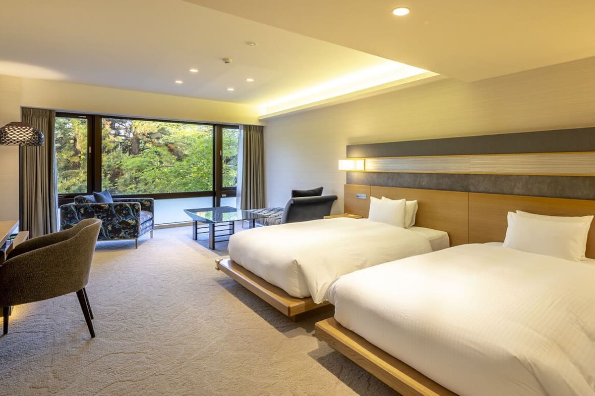 Une chambre d’hôtel avec deux lits, un bureau, une chaise et une grande fenêtre donnant sur un paysage verdoyant.
