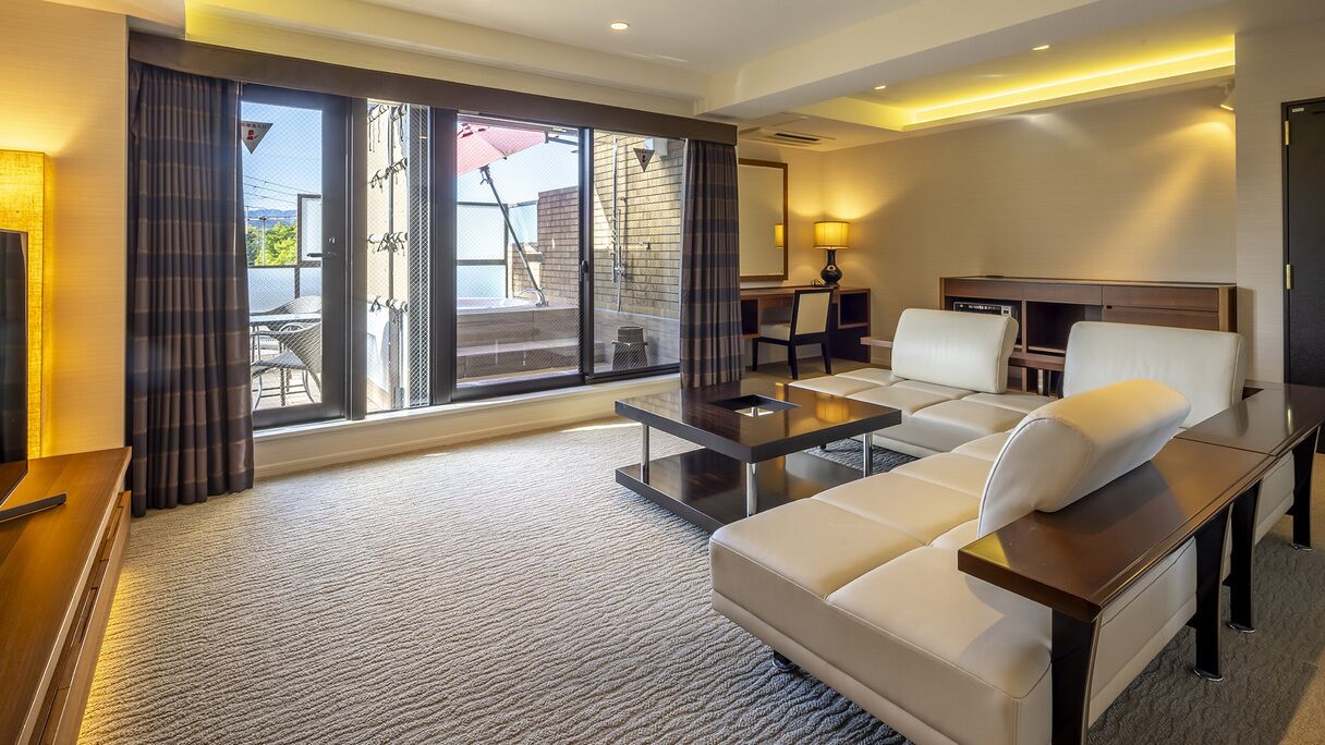 Ein Hotelzimmer mit Sofa, Couchtisch, Fernseher, Schreibtisch und einer großen Schiebeglastür, die zu einem Außenbereich führt.