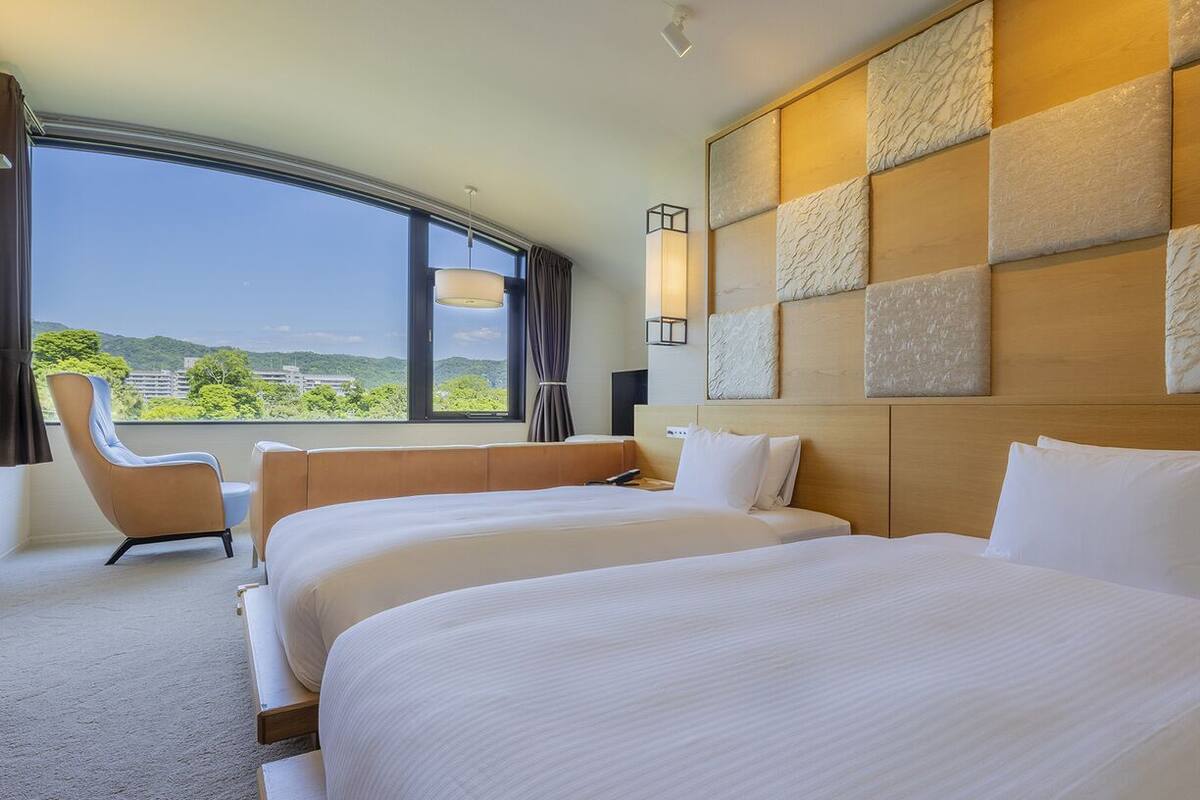 Une chambre d’hôtel avec deux lits, une grande fenêtre, une chaise et un panneau décoratif au mur.