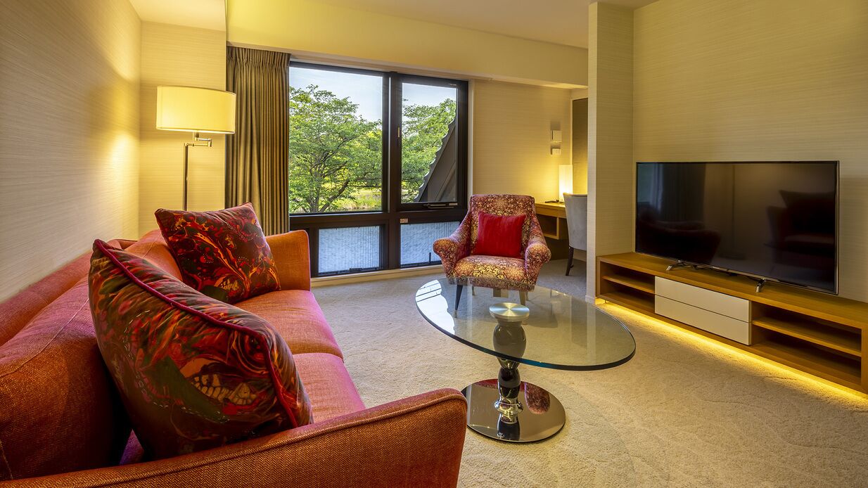 Ein Hotelzimmer mit Sofa, Sessel, Glastisch, Fernseher und großen Fenstern.