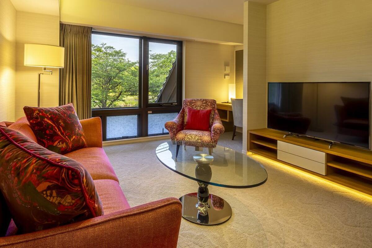 Une chambre d’hôtel équipée d’un canapé, d’un fauteuil, d’une table basse en verre, d’une télévision et de grandes fenêtres.