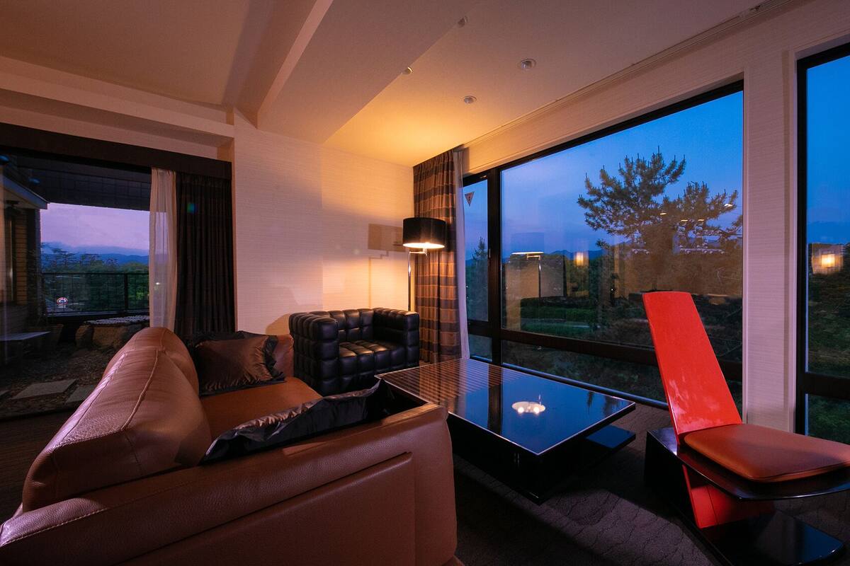 Un salon moderne avec un canapé, un fauteuil rouge, une table en verre et de grandes fenêtres donnant sur un paysage de arbres et de collines.