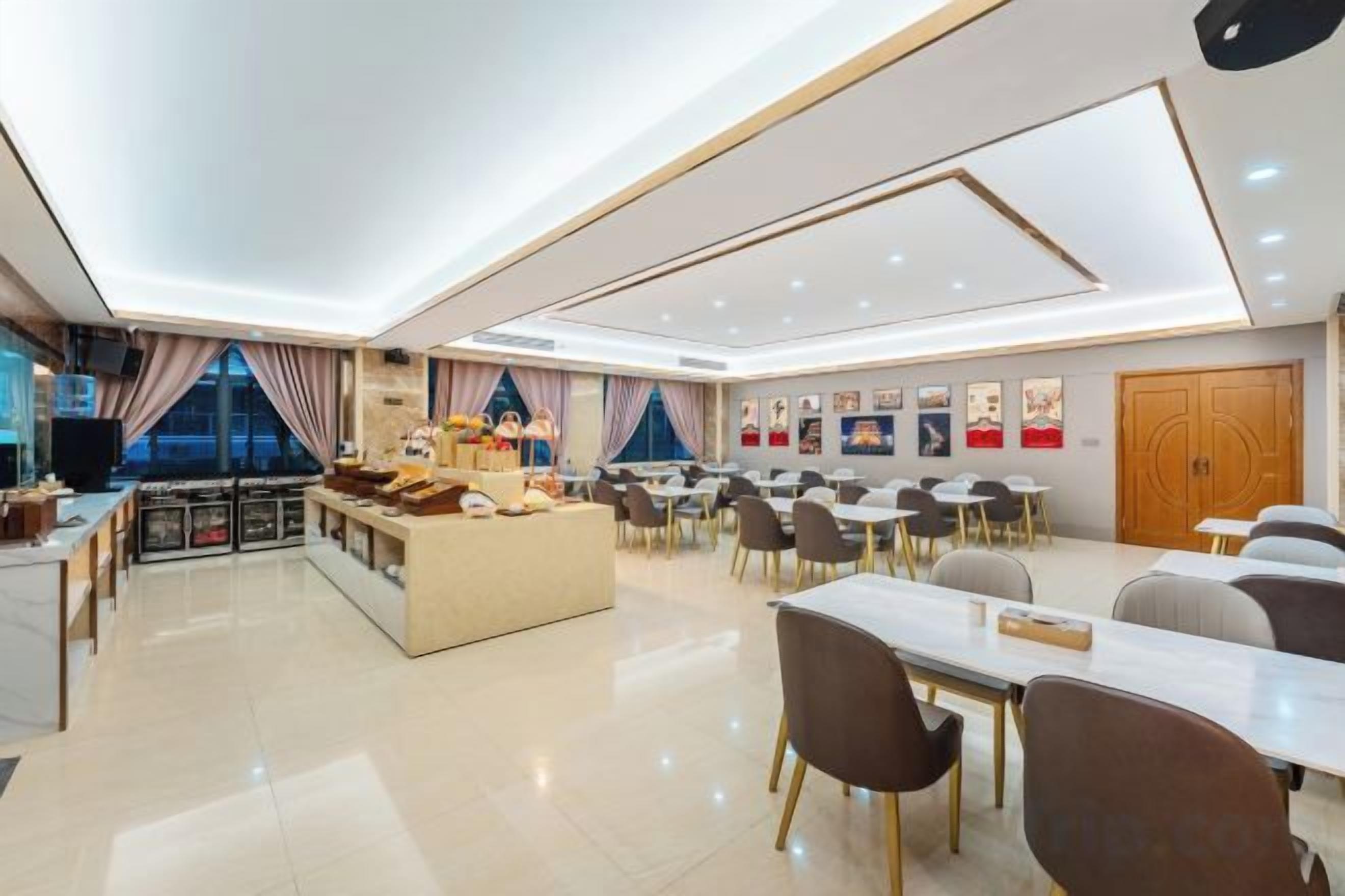 A spacious hotel dining area with multiple tables and chairs, a central seating area, and a kitchen visible in the background.
