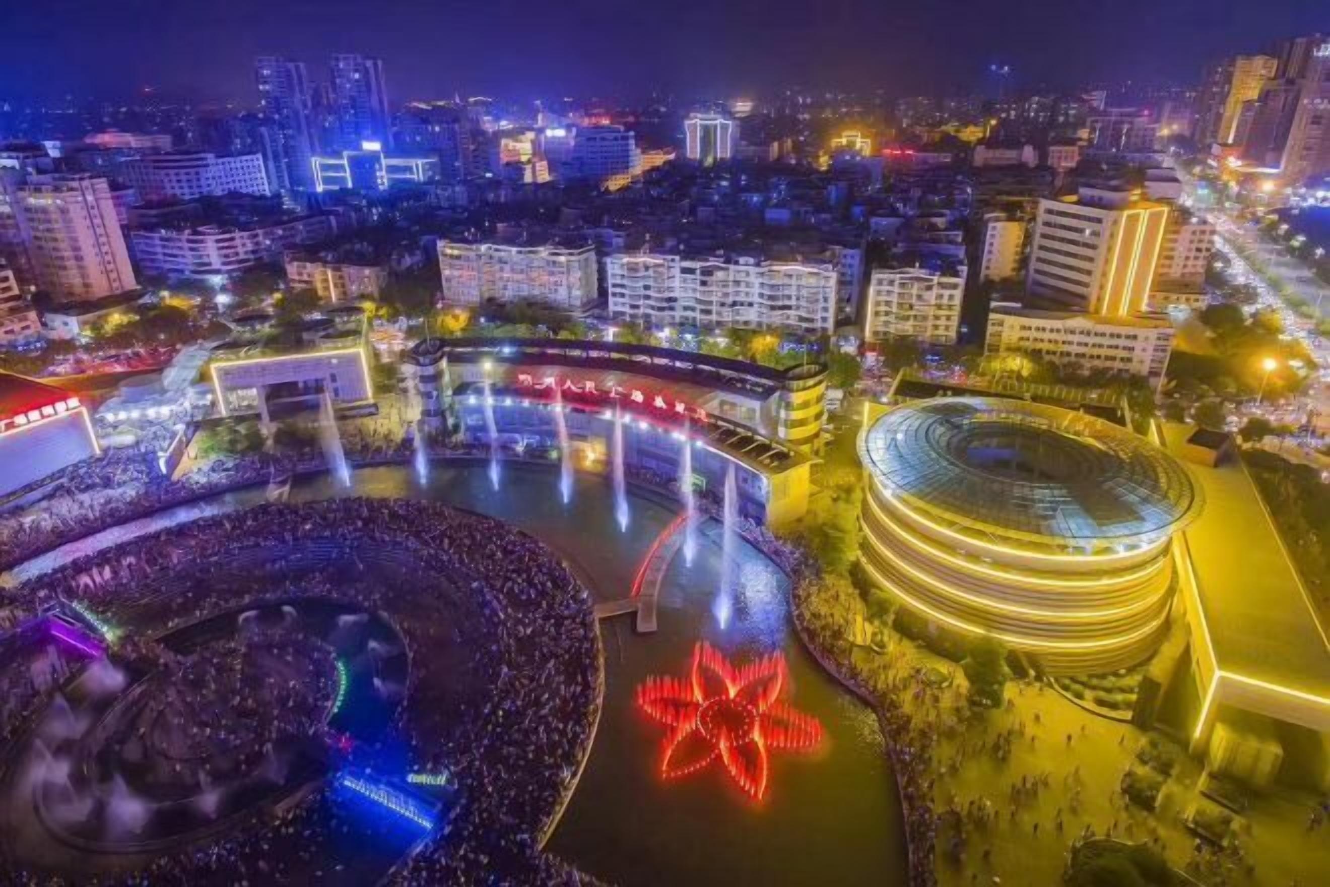 A nighttime cityscape with illuminated buildings, a large circular structure with a central fountain, and a crowd gathered around a brightly lit flower-shaped installation.