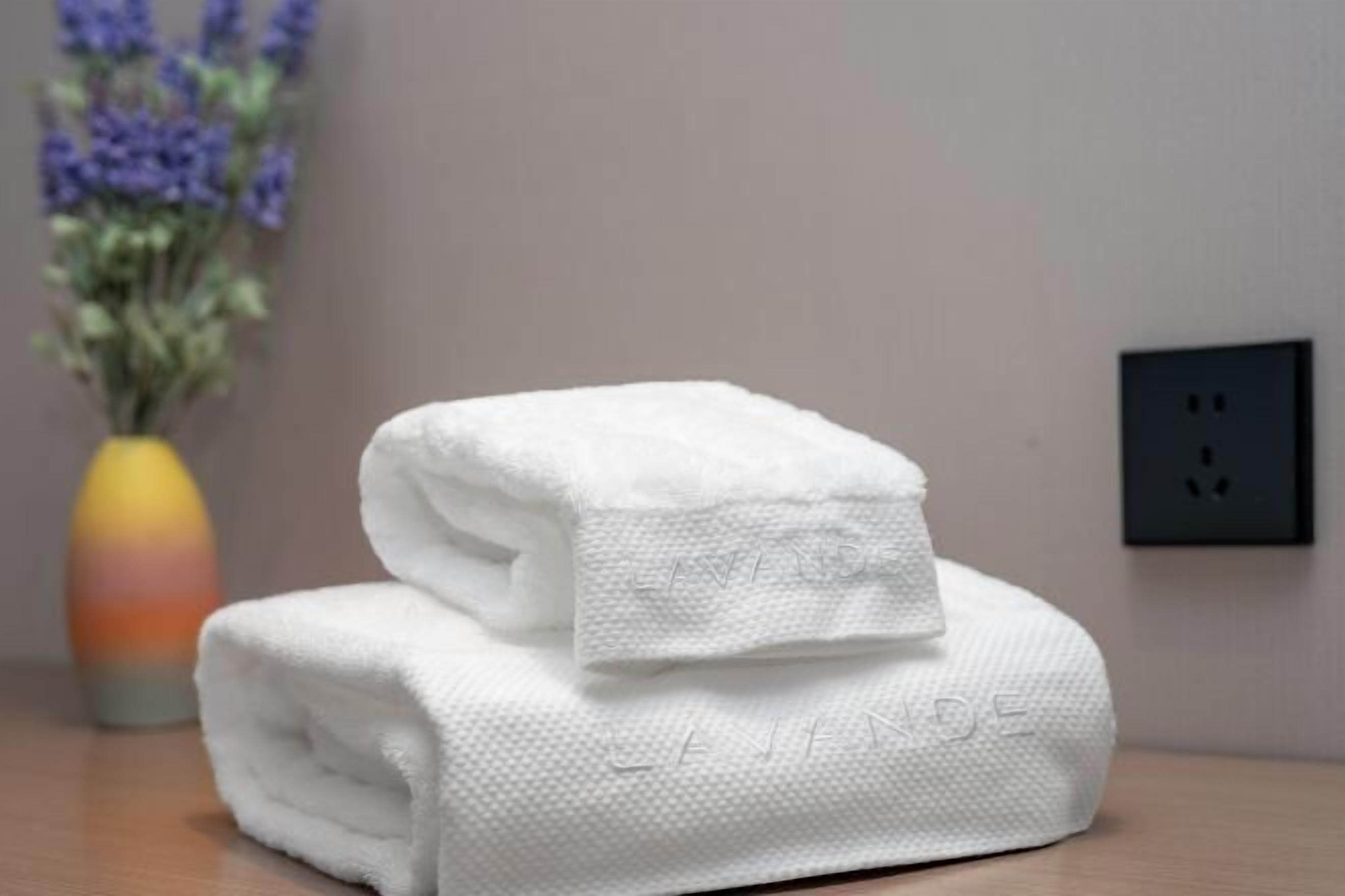 A neatly folded white towel with "LAVENDER" embossed on it, placed on a wooden surface with a vase of lavender flowers in the background.