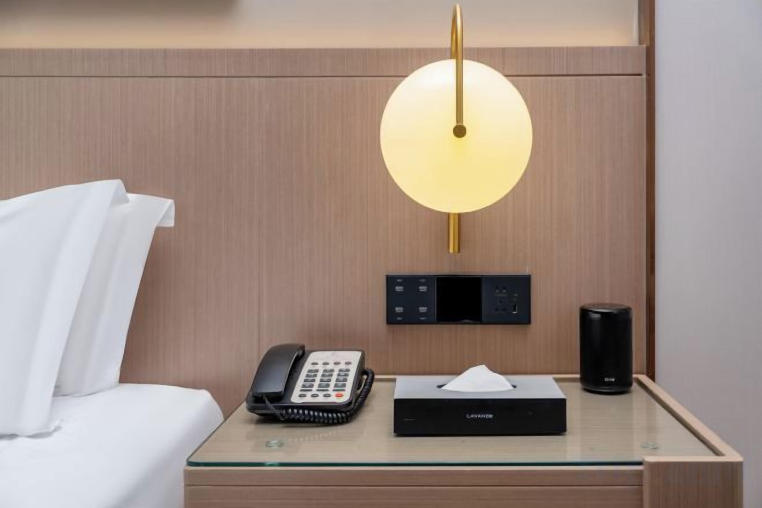 A hotel room with a bed, a bedside table with a telephone, a lamp, a box of tissues, and a control panel.