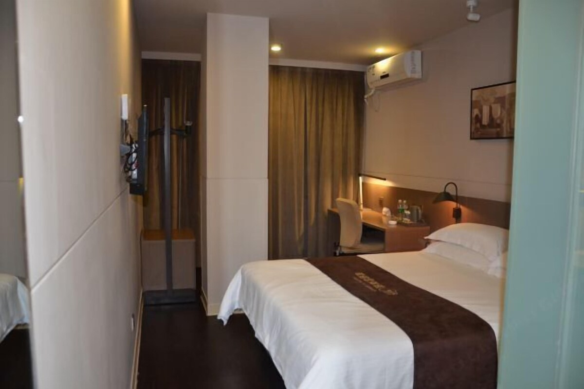A hotel room with a bed, a desk, a chair, a nightstand, and a wall-mounted air conditioner.