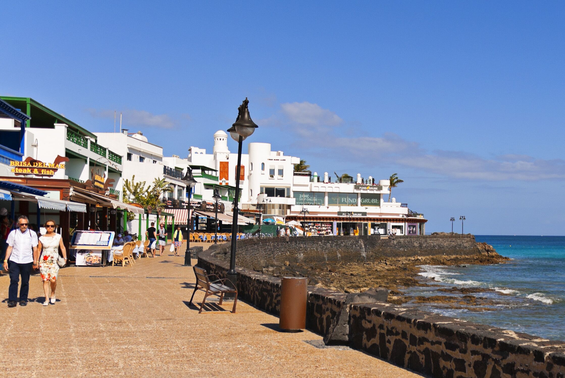 A coastal promenade with shops, a bench, and a trash can.