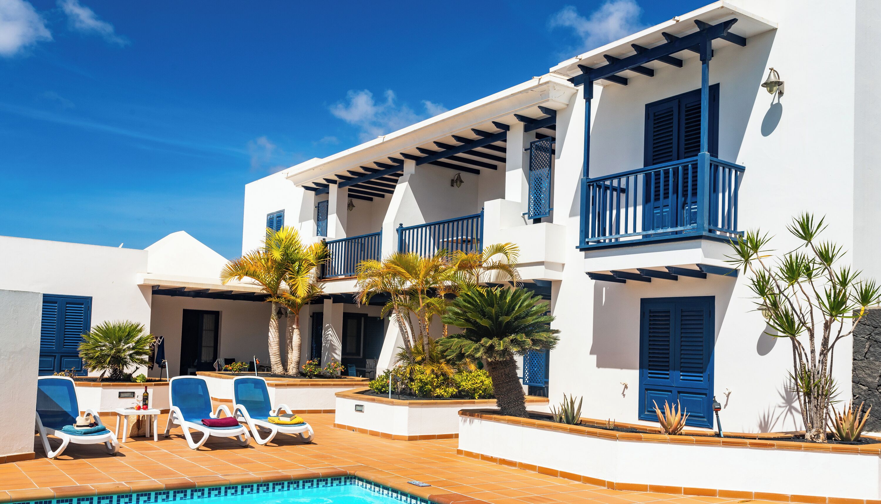 A two-story building with a pool, blue balconies, and tropical plants.