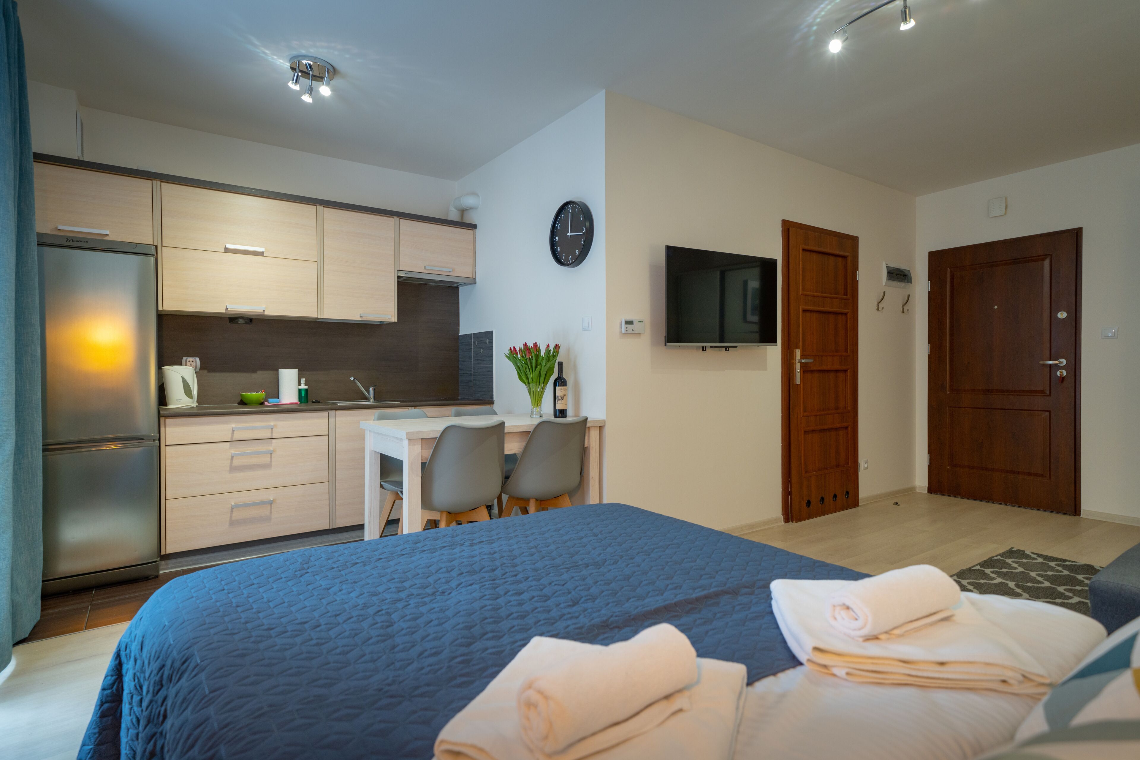 Een moderne hotelkamer met een bed, een bureau met stoelen, een kitchenette, een televisie en een klok aan de muur.